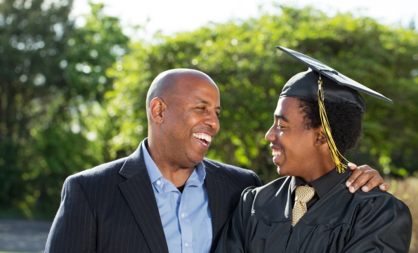 person in a blazer and button down shirt with their arm around a younger person (presumably their son) wearing a graduation cap and gown