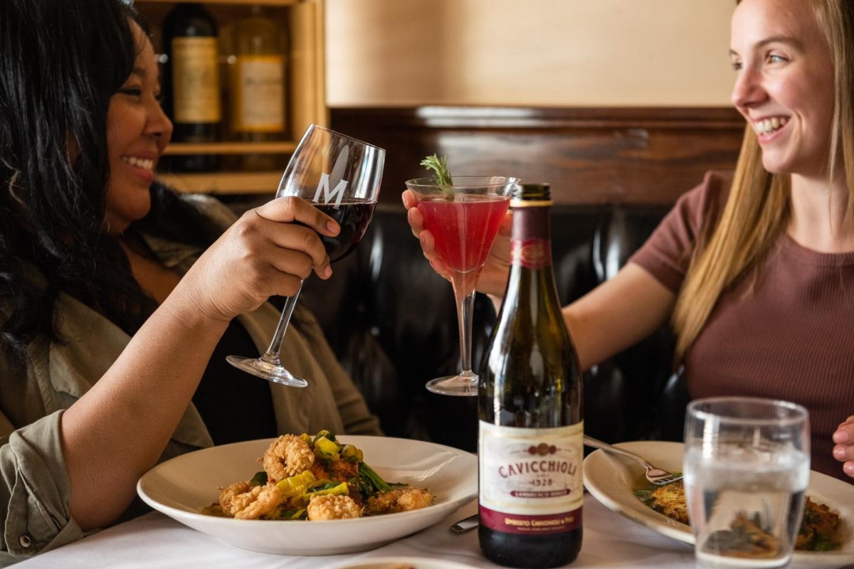two women cheers-ing wine glasses with plates of food in front of them