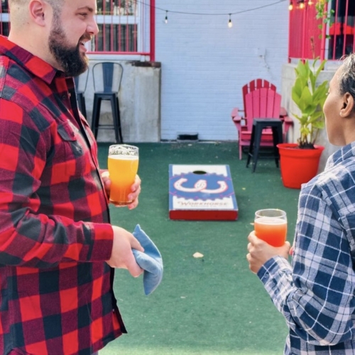 Two people holding beers and talking near a cornhole game