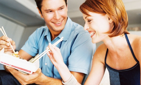 two people smiling and using chopsticks to get food from a takeout container